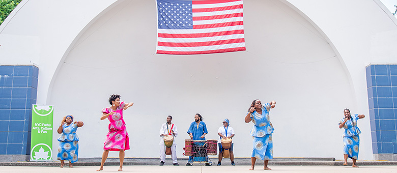 A group of women in traditional African wear dance on stage at a park the beat of the drums played by three men at the back of the stage.