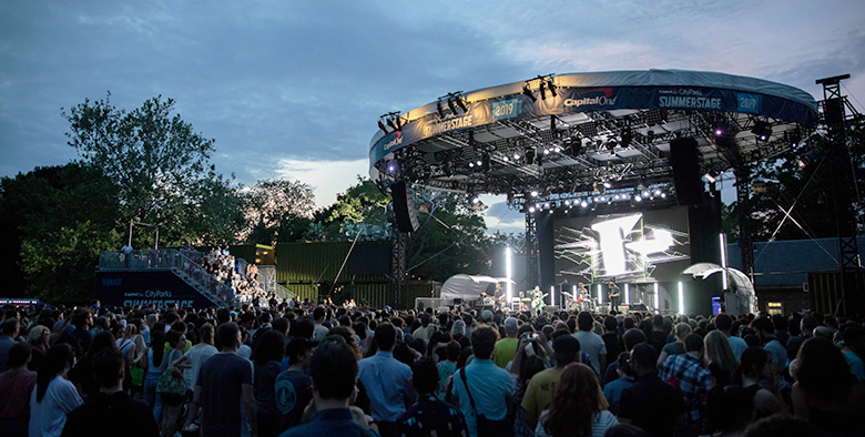 A crowd of people gather in front of a stage in a park for a concert.