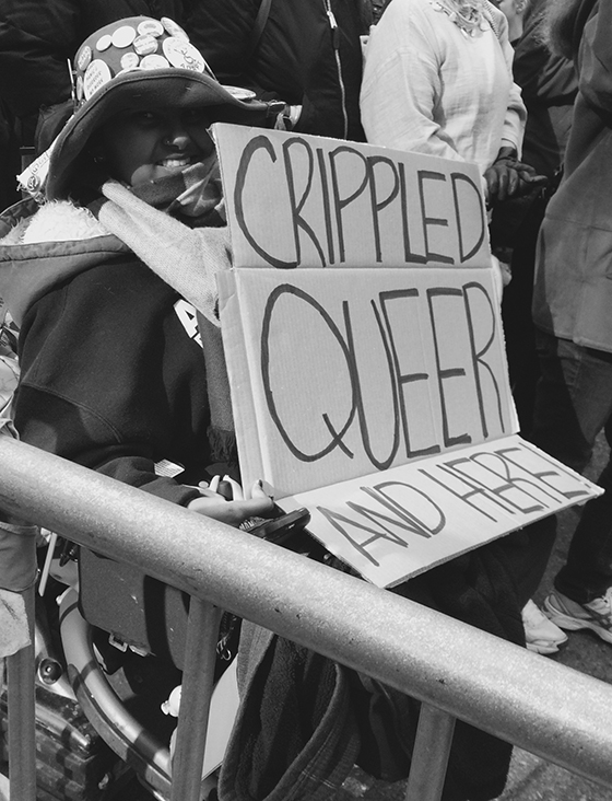 A woman in a wheelchair with buttons on her hat holds up a sign that reads "crippled, queer and here" while in the Women's March.