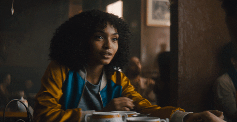A girl sitting at a table in a cafe nervously reaches out to touch her arm as she lovingly stares at her companion off screen and brushes his hands