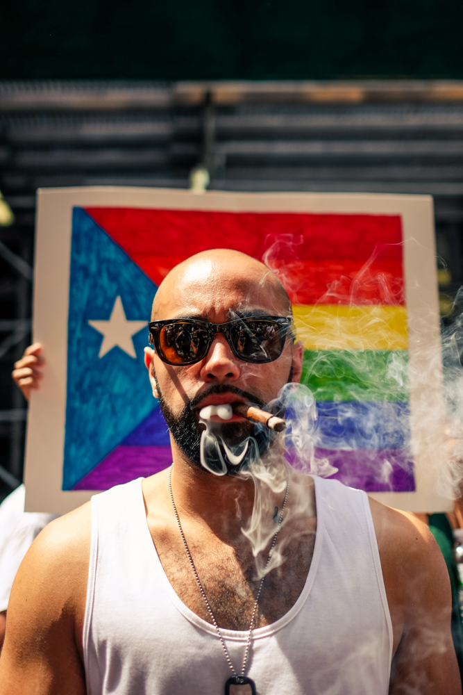 A man wearing sunglasses, a tank top, and dog chains, smokes a cigar while standing outside in front of a Puerto Rican flag in pride colors