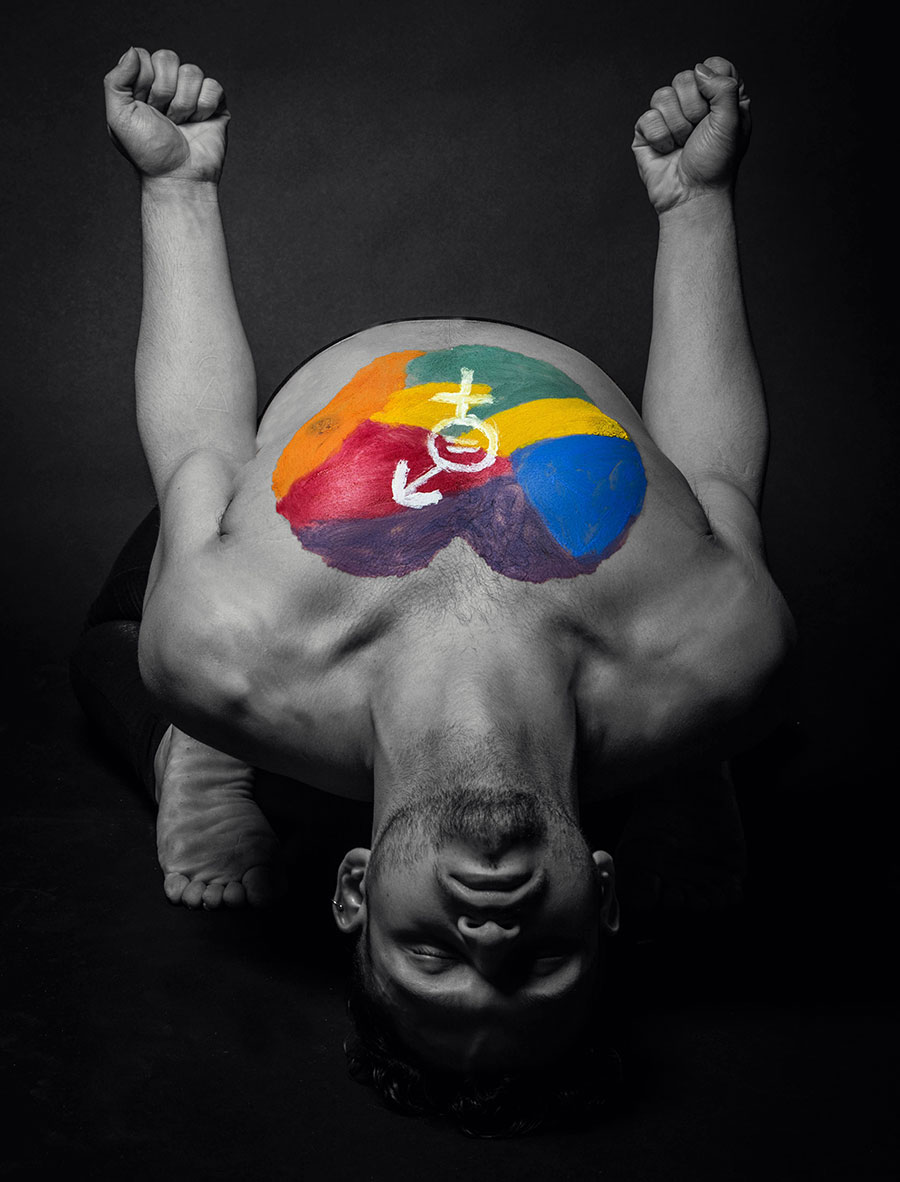 Black and white image of a person bent over, tummy up. A heart in pride colors with the gender equality sign in the middle is painted on the chest. , with fists in the air.