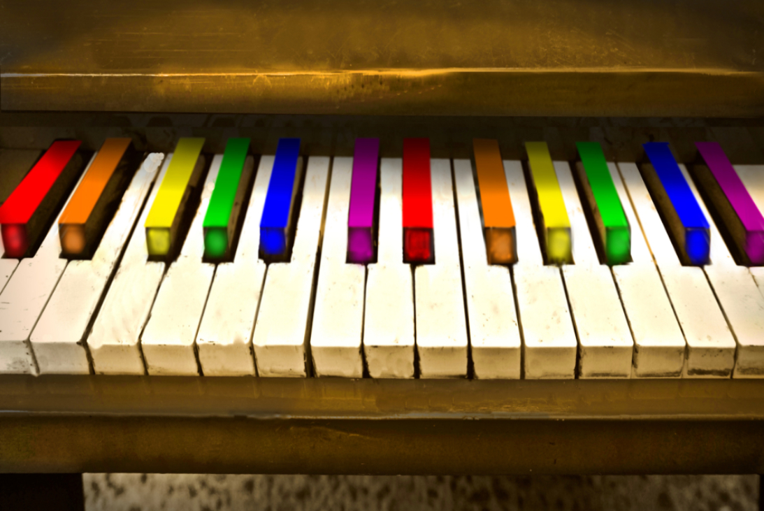 Image of a brown piano. The black keys have been replaced by pride colors.