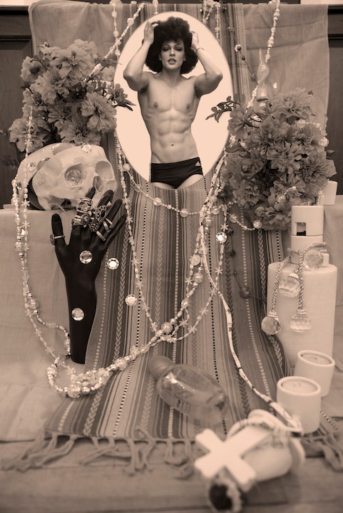 At the center of an altar with flowers, skulls, and jewelry is a person in a speedo playing in their afro while posing for a photo