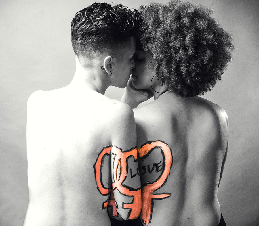 Black and white image of two people leaning in for a kiss with their backs turned to us. Their arms are connected by a painting of gender symbols in orange with the word love in black