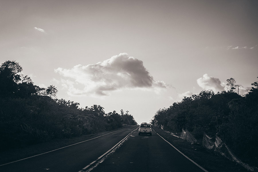 Black and white image of a car driving off into the distance on empty road surrounded by greenery and covered by an open sky with a few clouds