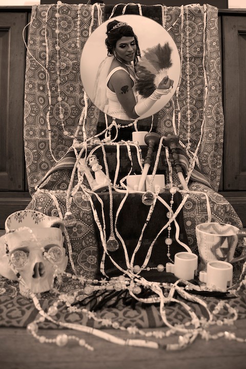 Circular framed image of a person dressed as a bride with feathers in hand on an altar decorated with skulls, dolls, jewelry, fabric and other keepsakes