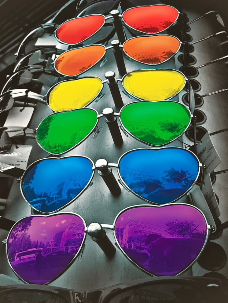 heart-shaped sunglasses in pride colors. Image of the photographer reflects in the shades