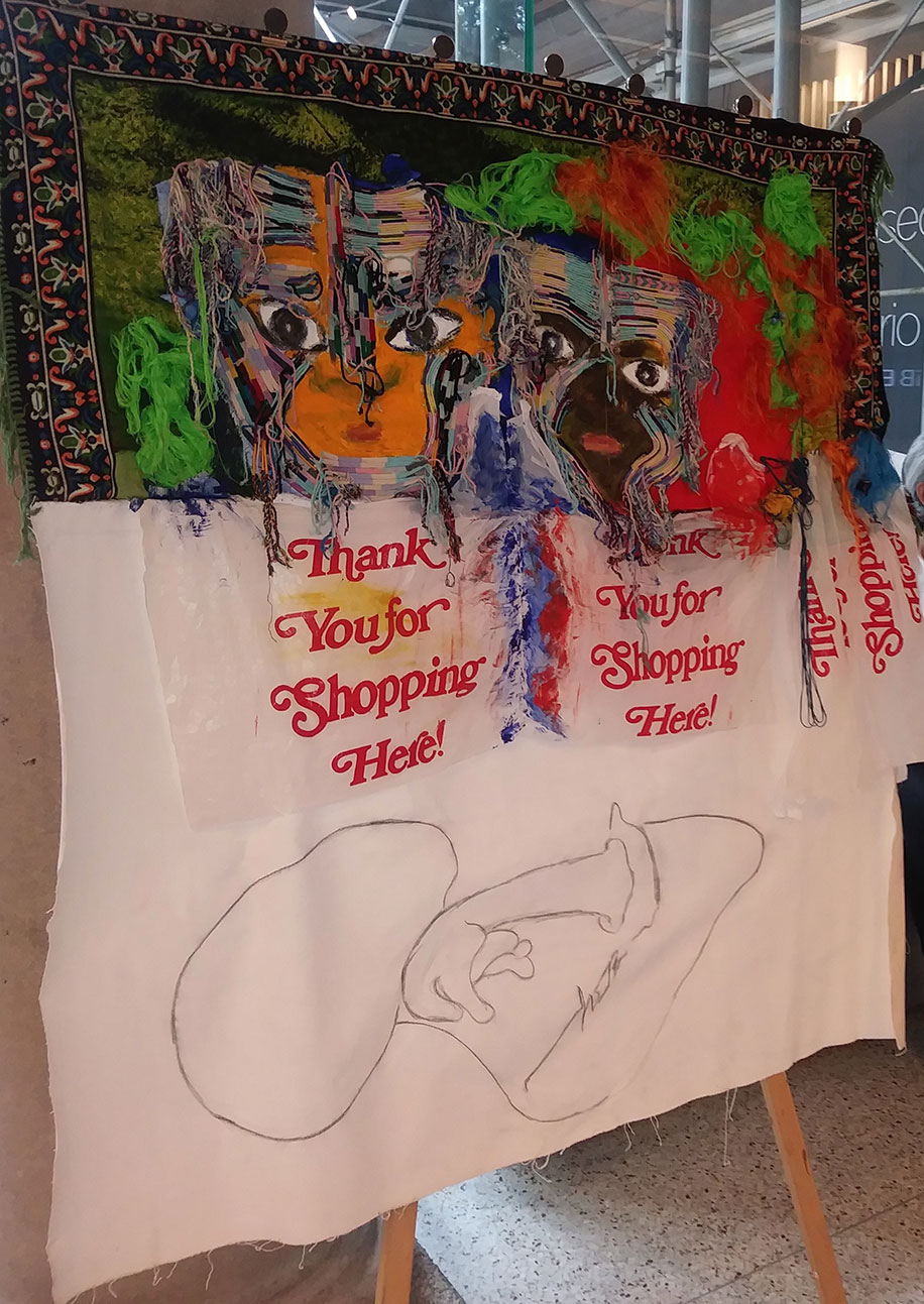 A hanging canvas of two faces, each above shopping bags that read Thank Your for Shopping Here; below the bags is an outline of someone propping their chin while lying down and looking up