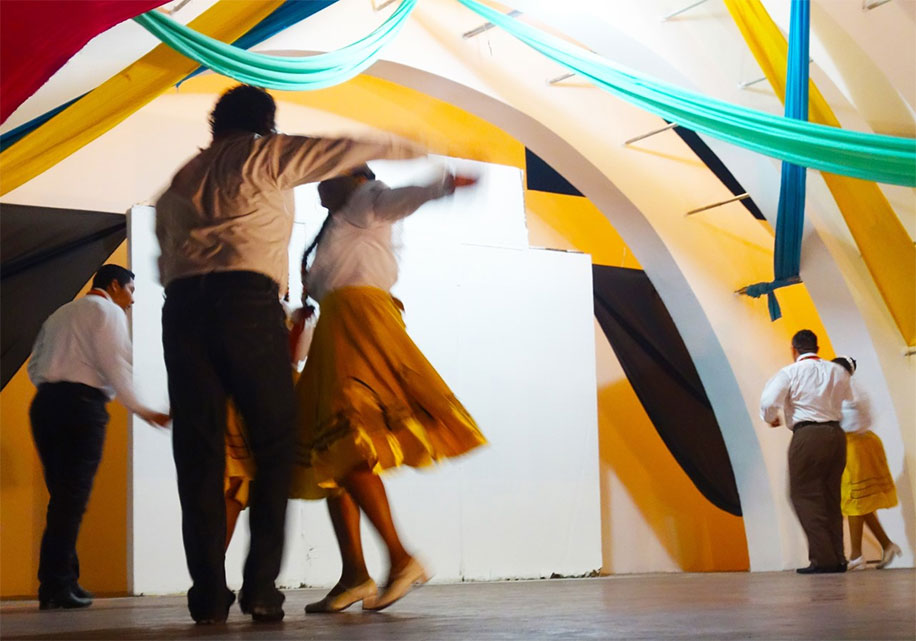 couples dance on a stage during a performance. The men are wearing white shirts and black pants and the woman wear long yellow skirts and white shirts