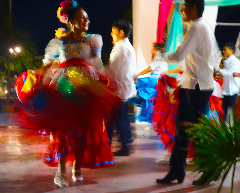 a man approaches a woman dressed in traditional wear, white shirt, a colorful tulle skirt, beads around her neck, and colorful flower-like hair decorations, as she dances on the dance floor