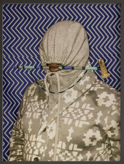 someone wears a gray hoodie with markings and holds a short staff with a golden tip and blue, aquamarine, and beige markings along the shaft; the person stands against against a blue and white chevron background