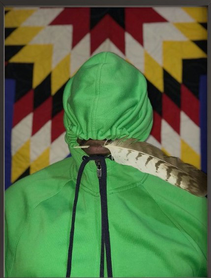 Someone wearing a neon green hoodie stands against a patterned background with yellow, white, red, blue, and maroon. The individual holds a feather in their mouth