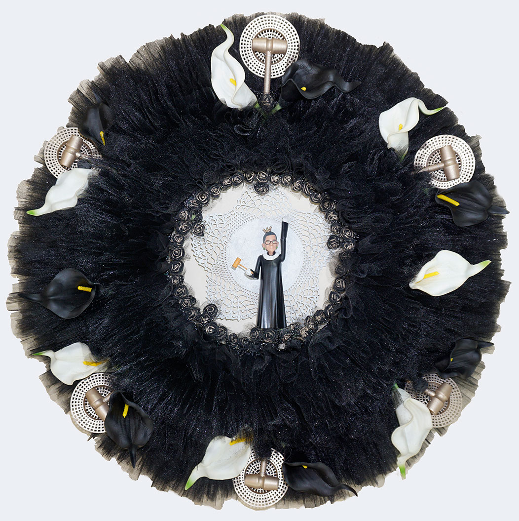 At the center of this wreath made of black tulle and decorated with white flowers and a judge’s gavel is an action model figure of Ruth Bader Ginsberg