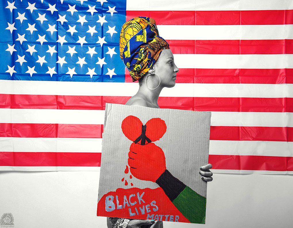 A woman in a headwrap stands sideways in front of the American flag. She is holding up a cardboard poster. The poster shows a red hand holding up a bleeding heart formed into a peace sign