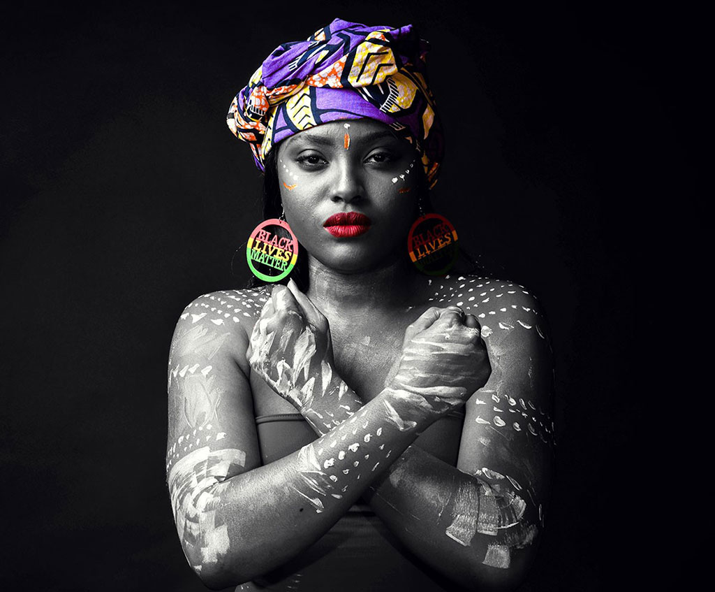 A woman wearing a headwrap and with tribal markings on her skin crosses her arms in front of her chest, fists clenched, engraved in her earrings are the words Black Lives Matter