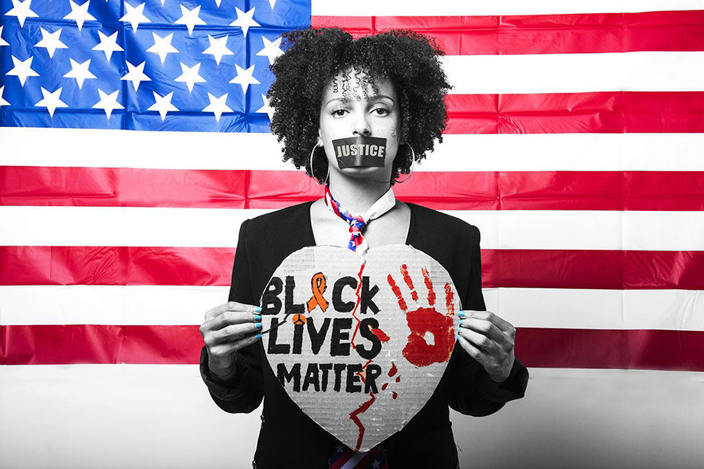 A woman in front of the American flag has tape labeled Justice covering her mouth, the American flag as a tie around her neck, and is holding up a broken heart sign that reads Black Lives Matter