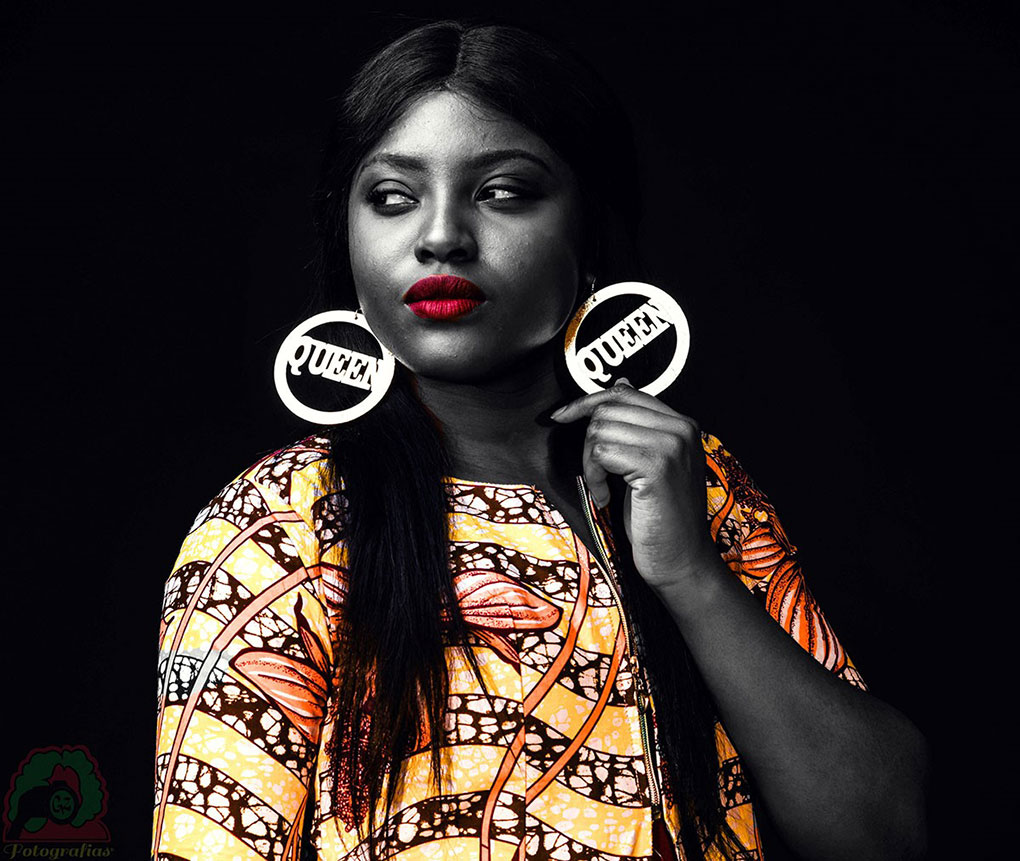 A woman hold on to one side of her earrings which has the word Queen