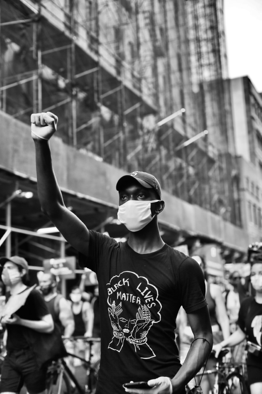 Black and white image of a protester, wearing a mask, a t-shirt that reads Black Lives Matter, and white gloves, puts his right fist in the air