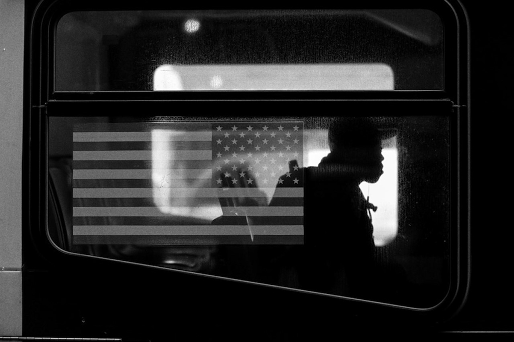 Black and white image of the American flag in the window of a train. The flag is flipped because it is displayed right-side from inside the train. A Black boy with a backpack and cap is sitting in the seats next to the flag