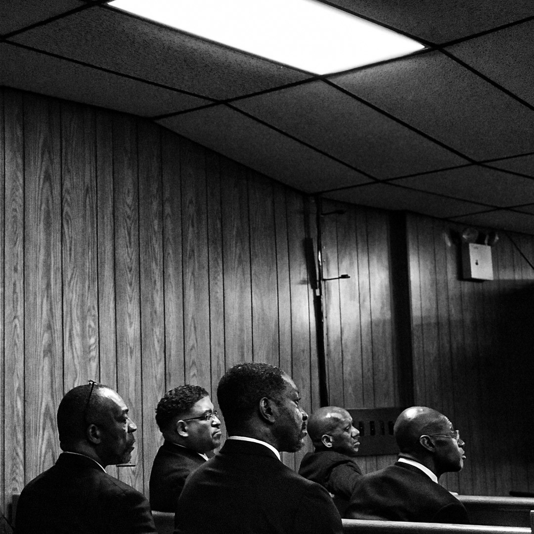 Black and white photo of a group of men in suits sitting together in rows of benches in a room. They appear to be looking to a speaker at the front of the room