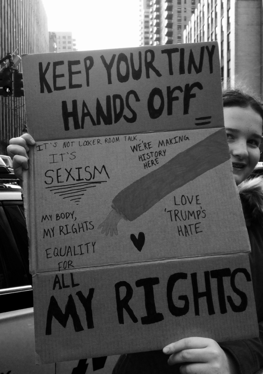 A protestor holds up a sign that reads Keep your tiny hands off, it's not my licker room talk, it's sexism, we're making history here, my body, my rights, love 'Trumps' hate, equality for all my rights