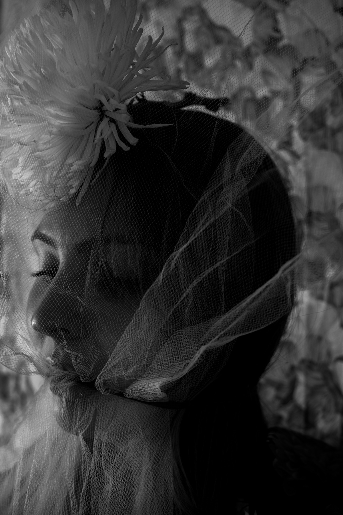 A woman with a flower in her hair closes her yes while under a veil-like netting fabric