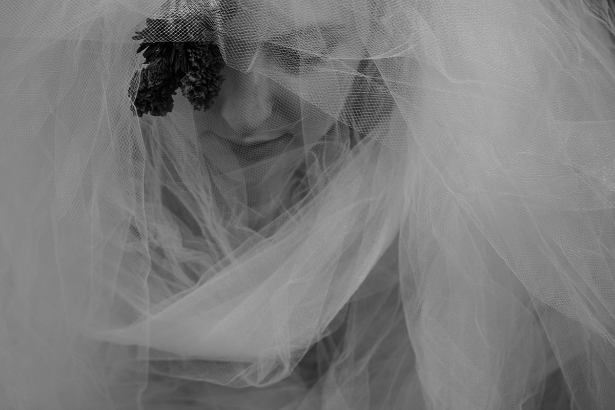 a woman with a flower in her hair and body beneath a veil-like fabric bows her head