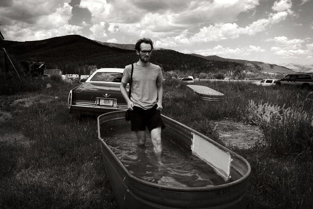 An individual stands in a boat of water placed in a grassy landscape where other boats and cars are parked or left abandoned