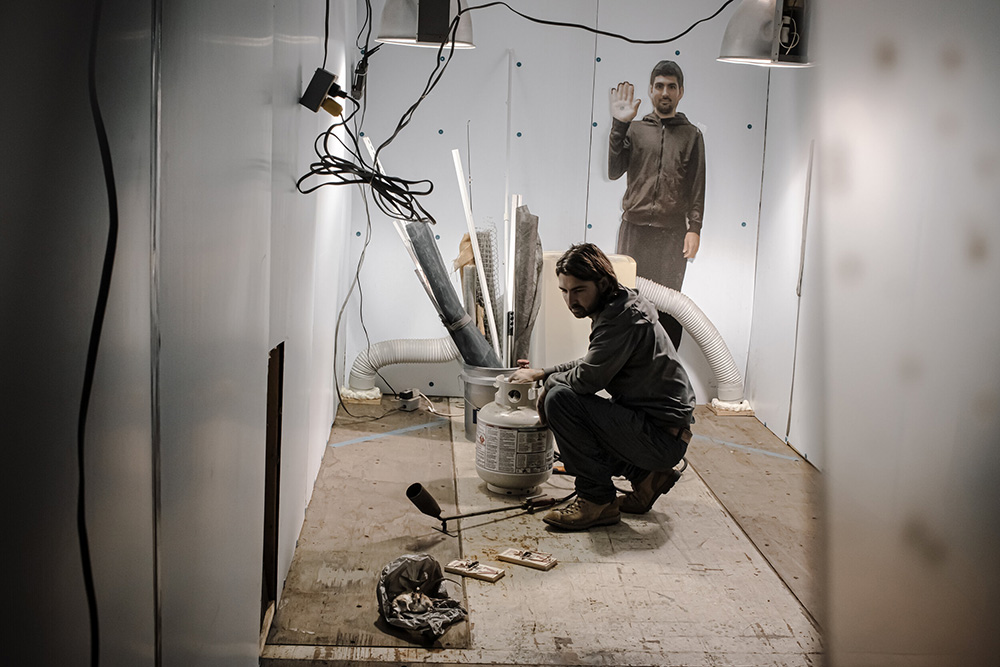 An individual works on materials in a room and there is an image of an individual holding up their hand as if to say hi on the wall