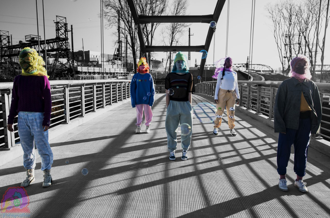 Individuals with fabric in various colors covering their faces stands in formation on a bridge