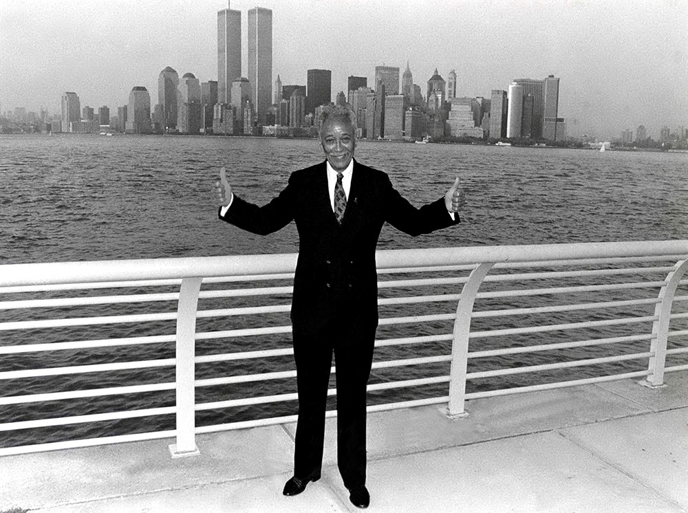 black and white photo of former Mayor David N. Dinks posing for a photo at the Brooklyn Promenade which features a view of the Twin Towers towering over the Manhattan skyline