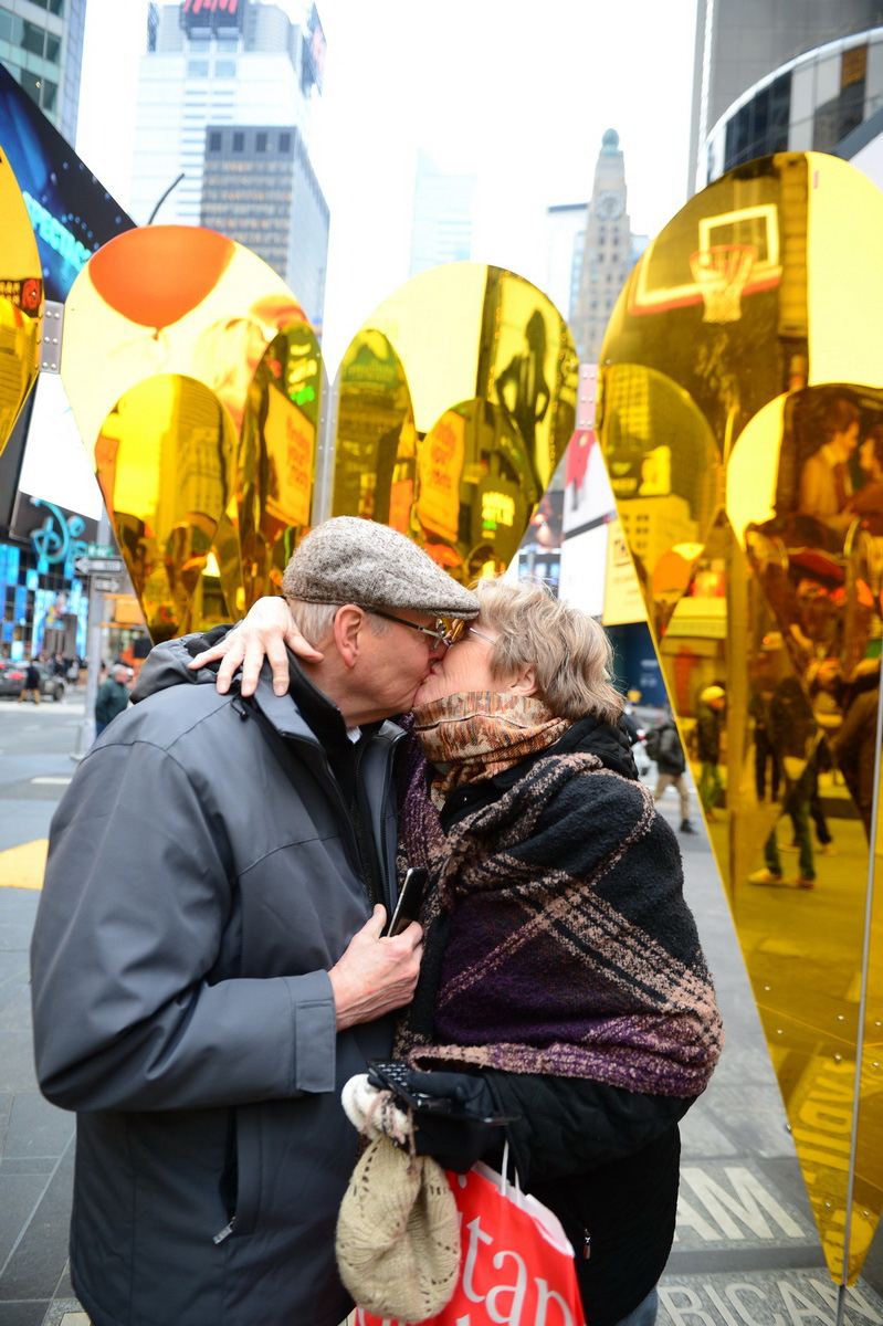an elderly couple kisses in front of an art exhibition of reflective hearts