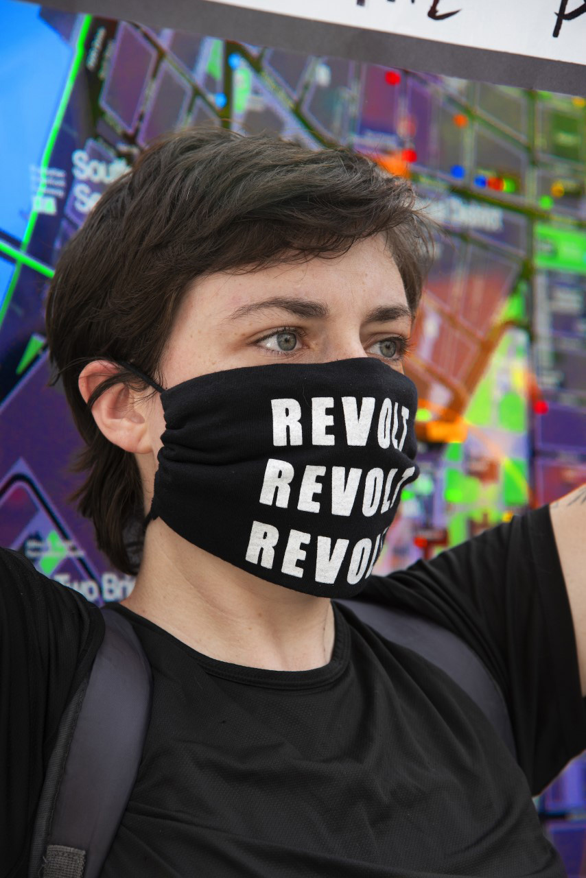 an individual holds up a Pride flag while wearing a mask that reads: Revolt. The word is repeated 3 times