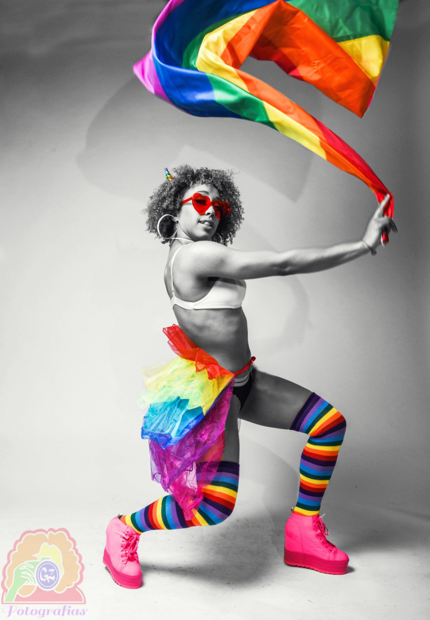Black and white image of a dancer wearing the following items in color: red sunglasses, pride-themed skirt, and pride-themed socks while waving the Pride flag