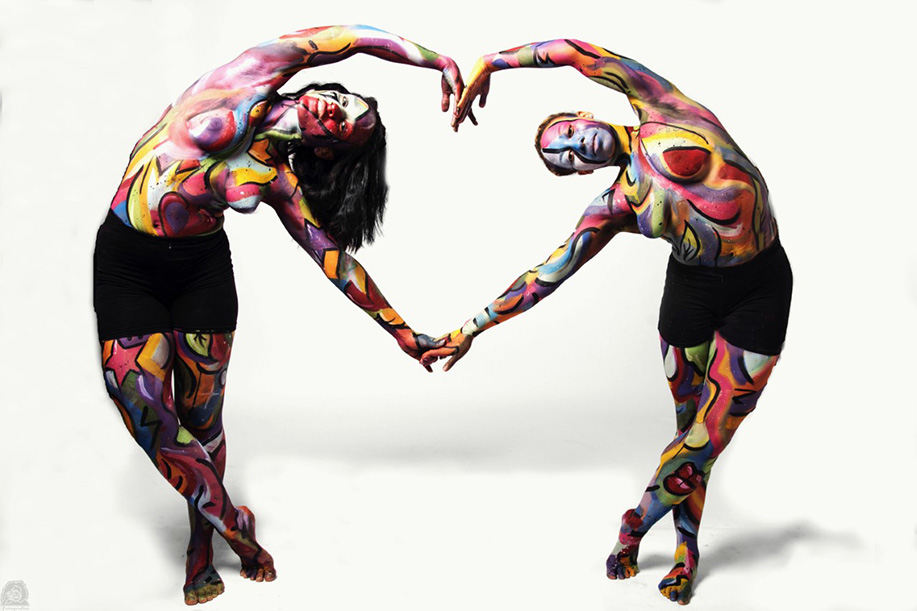 Dancers, their bodies painted in various colors, lean in to each other to form a heart with their arms