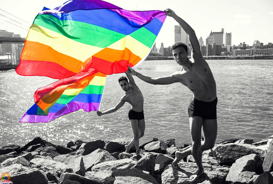 Two dancers hold up pride flags while standing on the rocks at the waterfront; the flags, the only part of the image in full color (the rest of the image is black and white) sway in the wind