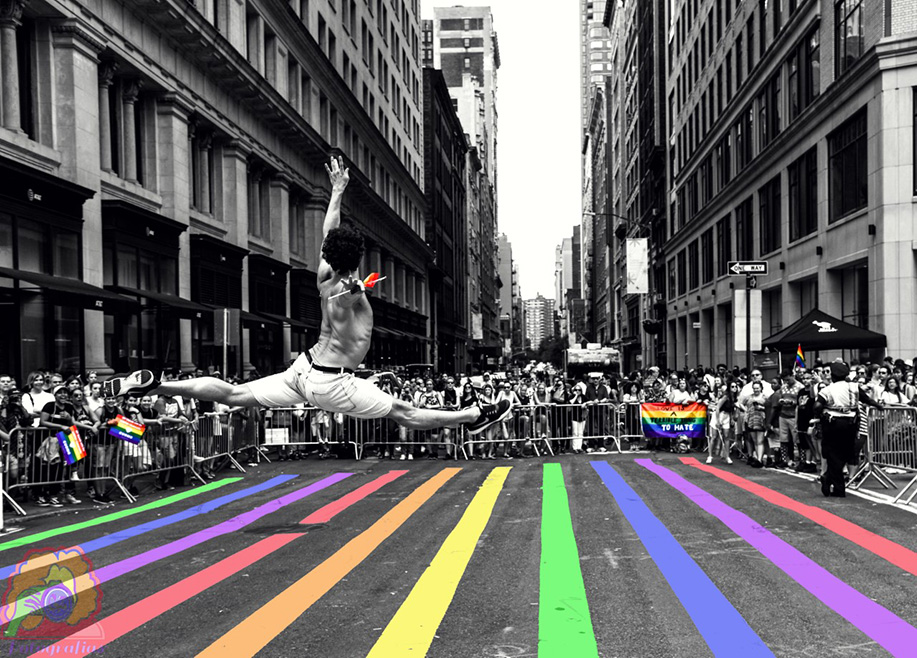 In this black and white photo, a dancer leaps above a sidewalk painted in pride colors while folks at the parade look on and show their pride flags. Only the flags and crosswalk are in color in this image