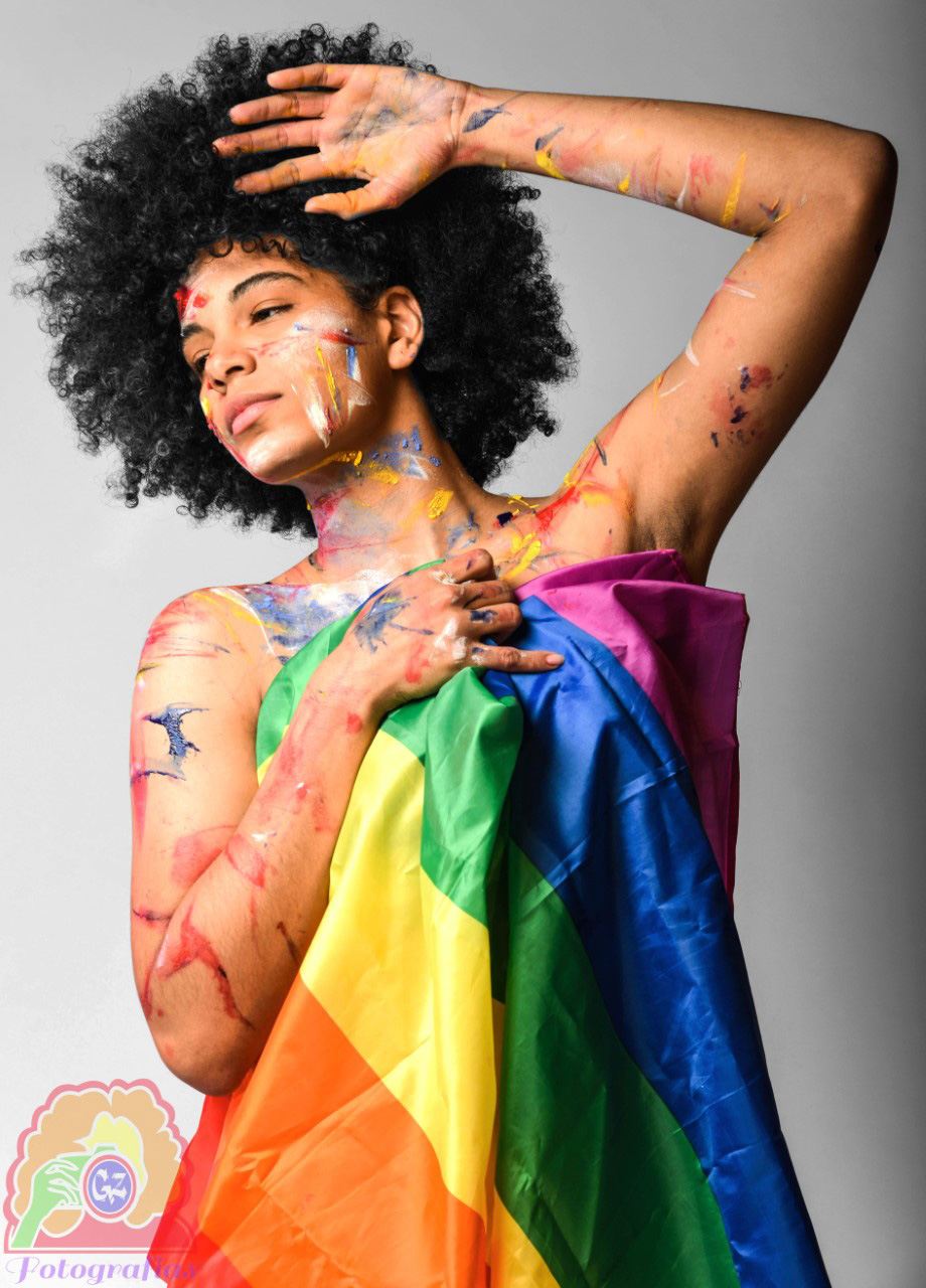 Image of a dancer looking away while covered in splashes of paint in pride colors and wearing the Pride flag