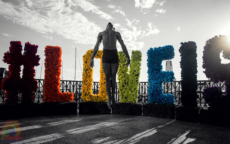 In this black and white image, a dancer leaps up into the air, and behind is a structure that reads #LOVEIS in color