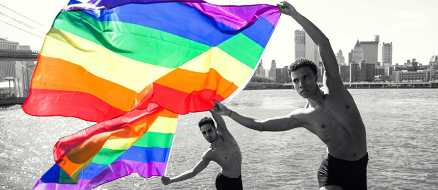 dancers fly the Pride flag while performing on rocks at the waterfront