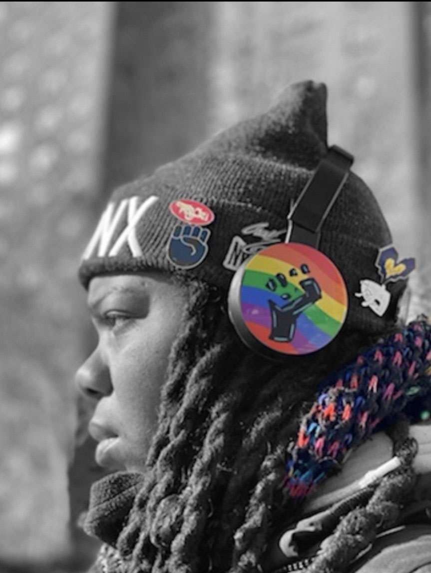 black and white image of an individual wearing over-the-ear headphones, the over-the-ear part is highlighted in rainbow colors with a Black fist, other fists and heart-shaped buttons on the individual's hat are also highlighted