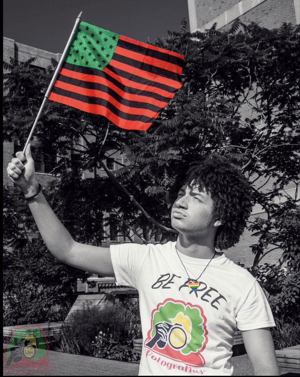 black and white photo of someone holding up the Pan African flag in color, also in color is the individual's t-shirt which features and individual with a fro taking a photo and the words: Be Free